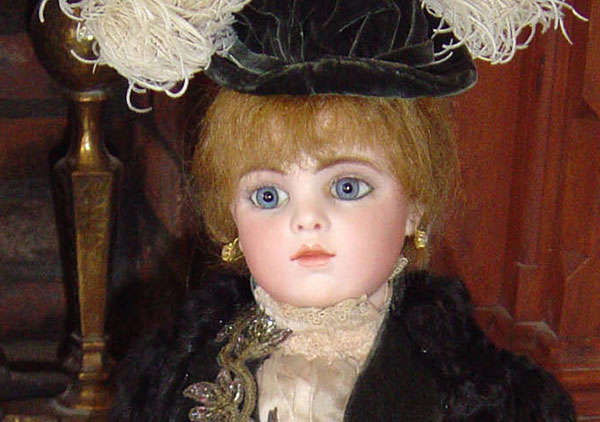 This close up photograph of the doll shows some finer details of the face, hat, and dress. Doll has Blonde hair and blue glass eyes. Her head shoulders, and forearm are bisque with upper arms & body thighs are kidskin leather. The lower legs and feet are celluloid. Tiffany mounted, fake pearl pierced earrings hand from her pierced ears. She is wearing a late nineteenth century style black dress with a feathered hat.