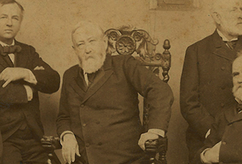 An old sepia-toned photo featuring Benjamin Harrison and his cabinet. Harrison leans forward in a decorative wooden chair, propping himself on the arm rest with his left arm.