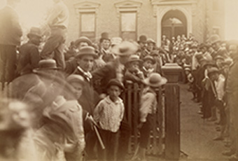 Photograph of crowd in front of Harrison's North Delaware Street home in 1888. Benjamin Harrison is standing near the front door slighlty to the left. Image shows the front of the house as it looked in 1888, picket fence at front sidewalk, and crowd of mostly men and young boys. A few women stand behind Harrison at the front door. The crowd is lined up on the rightside of the walk leading to the front door, in the yard to the left, and on the front sidewalk to the left.