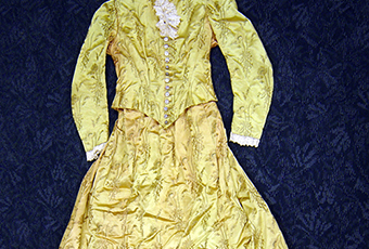 Gold flowered silk one piece dress marked as being Caroline Harrison's. Fabric is a gold color and different shades outlined in black of a design of long stemmed flowers. Like a poppy or daisy. Lace trimmed collar and cuffs. Lined, bottom edge of skirt inside small ruffle of a stiff netting. 13 buttons on front of bodice. Bodice attached at back with skirt. Metal stays some coming through fabric.