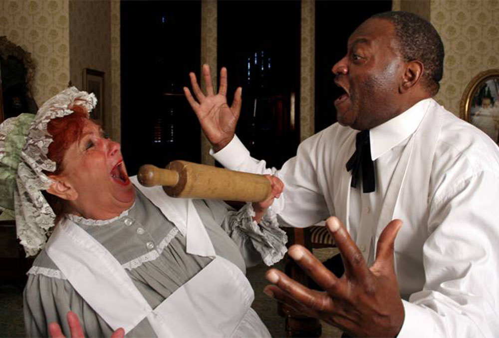Photograph of two candlelight theatre actors in the middle of a performance. On the right is a male actor, screaming in agony as a rolling pin protrudes from his bleeding arm. On the left is a woman wearing a bonnet and apron who seems shocked at the gory sight.