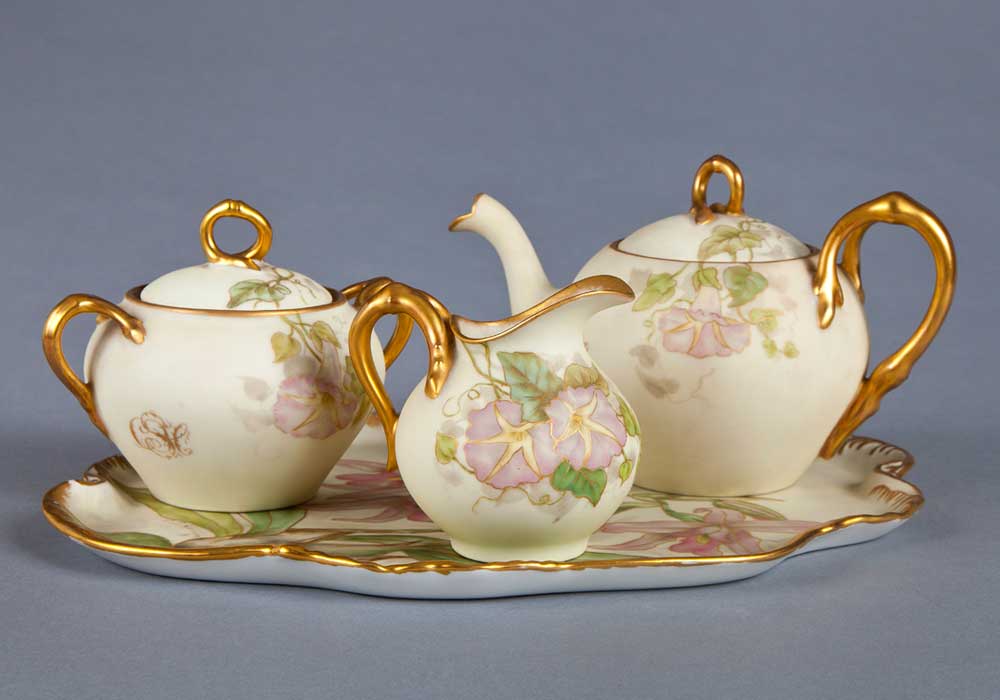 Photograph of luxury china set. The set includes a gold lined tray with a floral pattern, complete with a matching kettle and two small jugs.