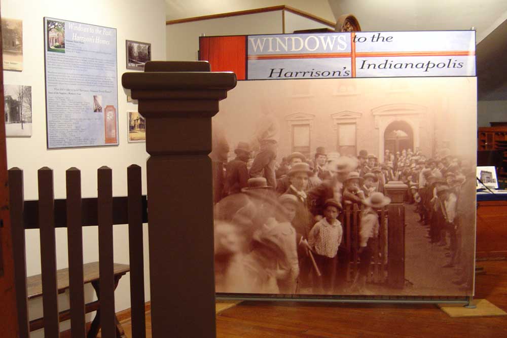 a photograph from the third floor of the benjamin harrison home, showing a railing and some photography from the eighteen hundreds of a large crowd gathered outside the presidential home.