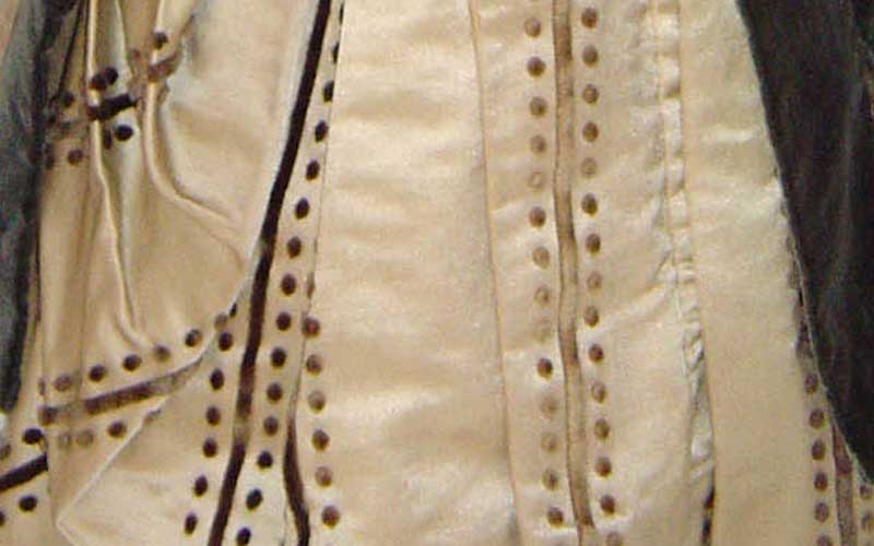 Photograph showing finer details on the dress skirt. There skirt is ornately connected with buttons and lining the accents vertically, in gold. The fabric is lightly wrinkled.