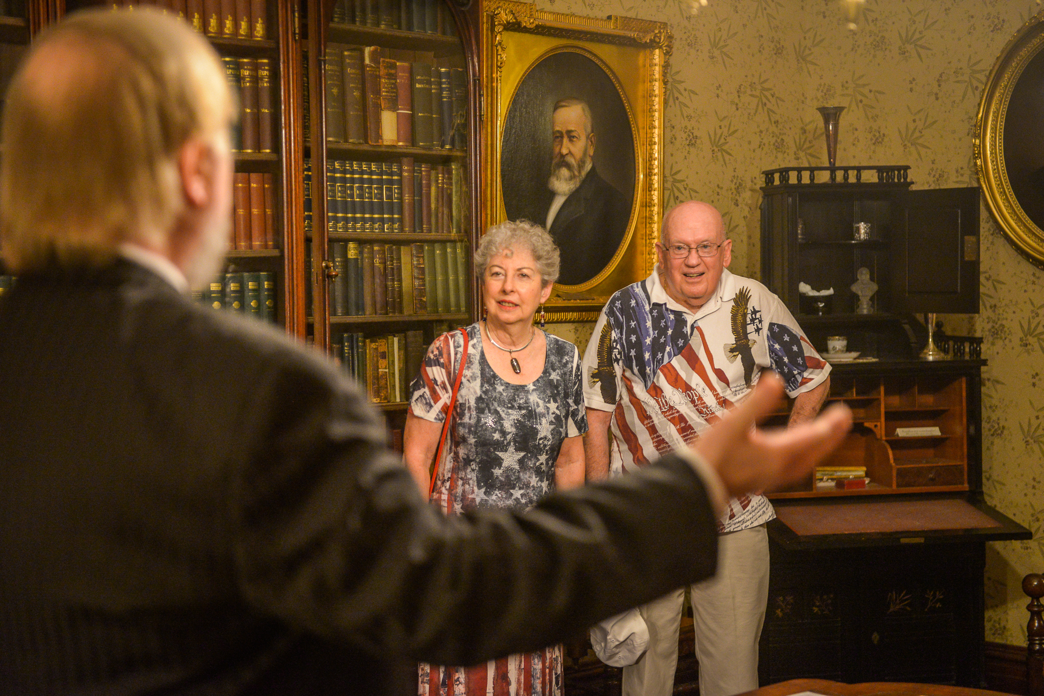 Photograph of a senior couple in benjamin harrisons office, both wearing shirts with american flag imagery. In the foreground can be seen a man with open arms.
