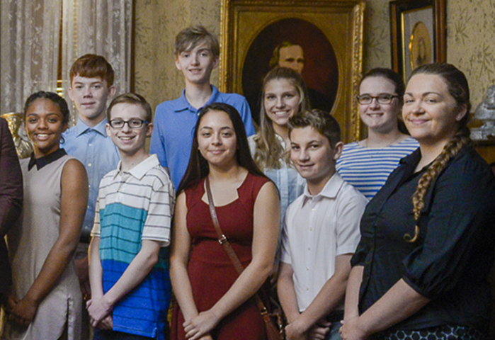 Photograph of students in the Harrison library. The photo features nine students standing with their hands formally folded at the front.