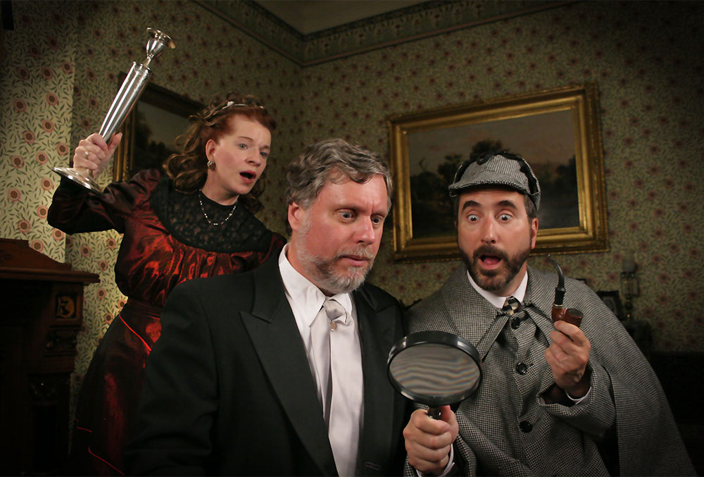 Photo of three candlelight theatre actors in the middle of a performance. The photo depicts Sherlock holmes and Watson in a dark room. Holmes holds a pipe in his hand, and wears an expression of surprise as watson looks onward with a magnifying glass. Unbeknownst to them, there is a woman dressed in red behind them with a candlestick, preparing to attack them.