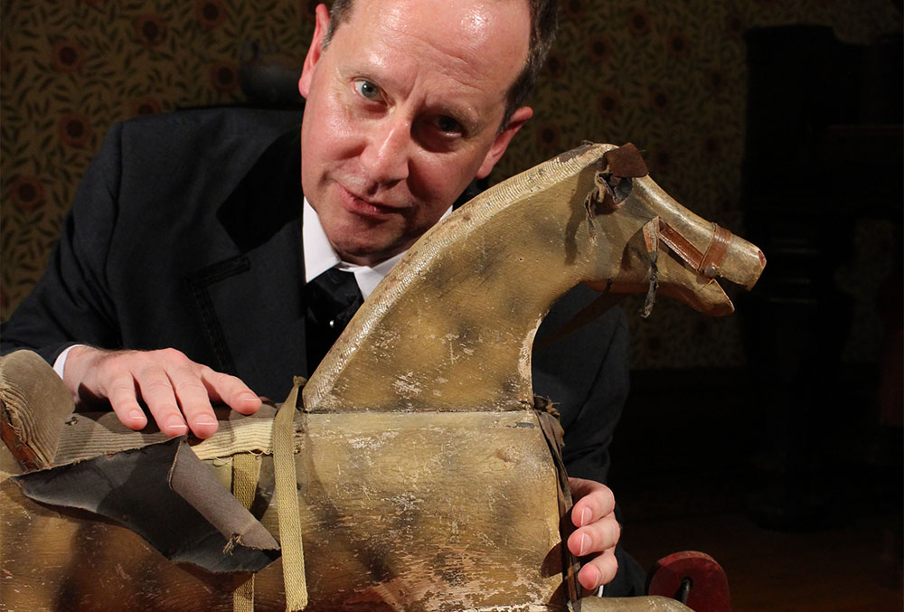 Photograph of a candlelight theatre actor. The character leans over a children's horse rocker toy, with one hand on its saddle and one hand on its chest.