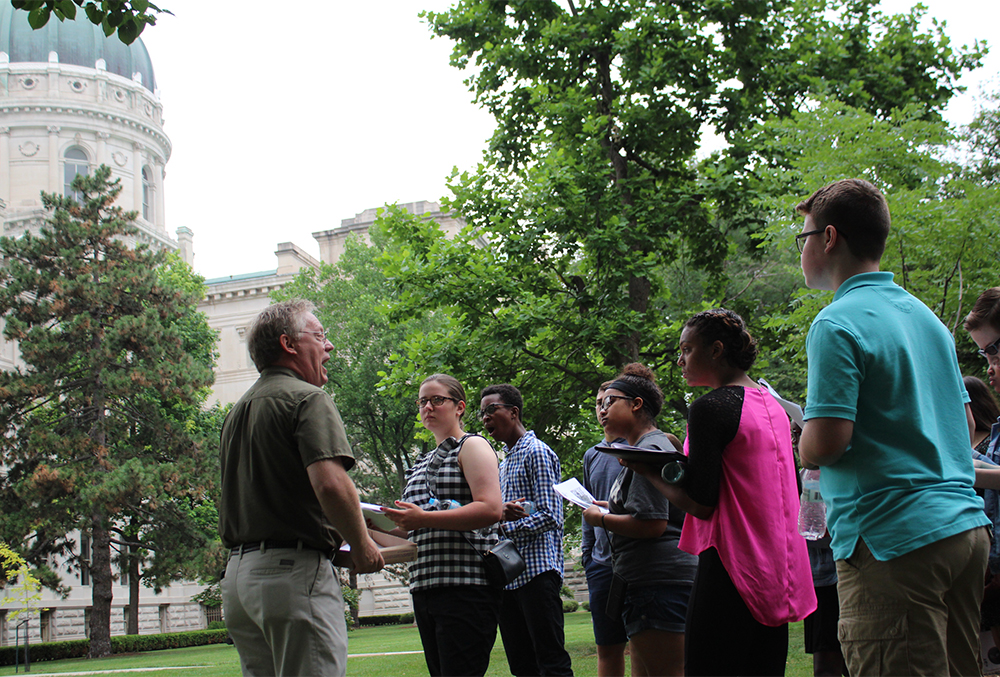 Photo of a small crowd of people having a discussion near the statehouse during the summer. They are surrounded by leafy green trees.