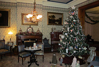 Photograph of a room in the benjamin harrison home, with a tree festively decorated and featuring presents. The room itself has a small table with chairs, adorned with a tea set. The walls have a floral pattern, several paintings, and a chandelier.