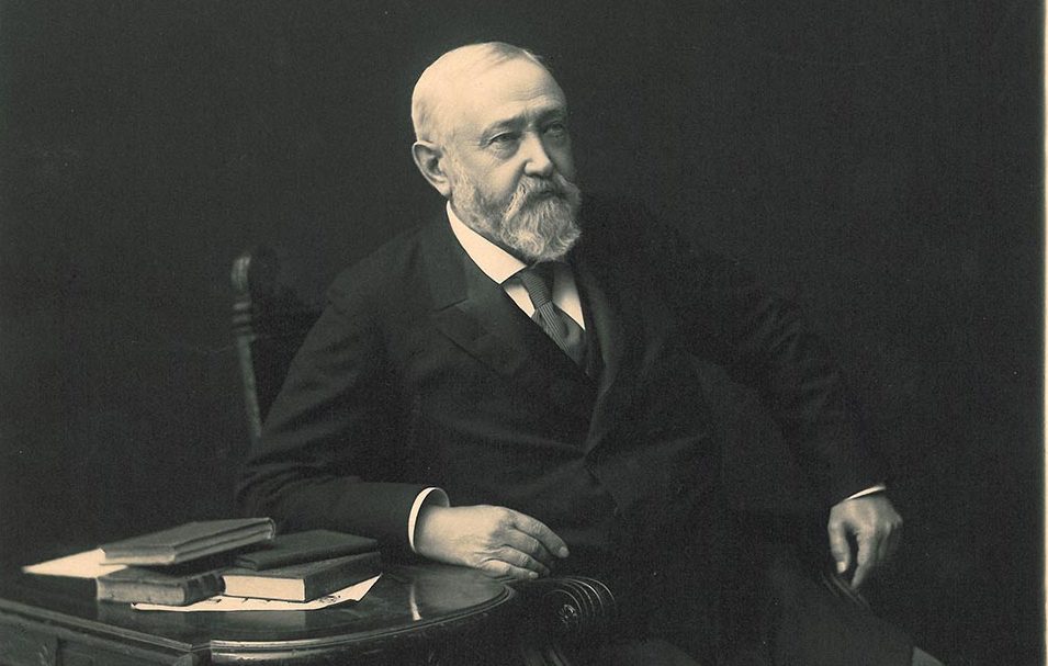 Photographic image of Benjamin Harrison sitting in a chair. He leans one arm against a side table next to him with four books haphazardly stacked on top of some loose paper. He is brightly lit in front of a dark background.