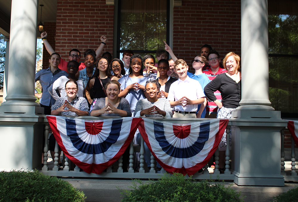 Photograph of a group of people cheerily gathered for a photo on the porch of the benjamin harrison home. The photo contains around twenty people, some of which are throwing their arms up in celebration.