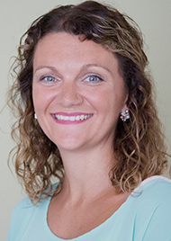 Portrait of Heather Wing, a middle aged woman with dark blonde hair. She has light blue eyes, small earrings, a light blue sweater, and smiles jovially.