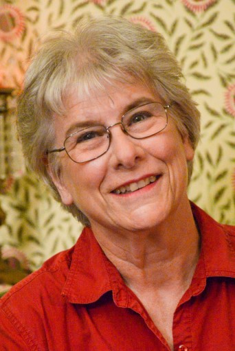 Photo of Jill Whelan, an elderly woman with short platinum blonde hair and ovular glasses. She wears a red dress shirt and smiles in front of a floral patterned wallpaper.