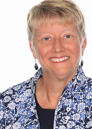 Photo of Jan Viehweg. A middle aged woman, she poses against a white background. She has short blonde hair, blue eyes, and wears a blue floral patterned shirt, droopy earrings, and a joyous smile.