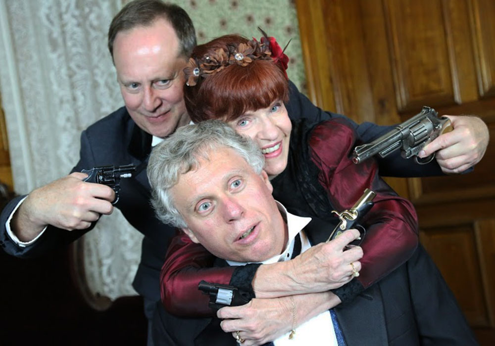 Photo of three candlelight theater actors having a sort of mexican standoff. Each actor appears to have another in a hostage situation, pointing guns at each other.
