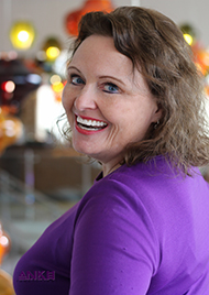 Portrait of Judy Lombardo, a middle aged woman wearing a purple cardigan. She looks over her shoulder with a big gleeful smile. She has brown hair and blue eyes.